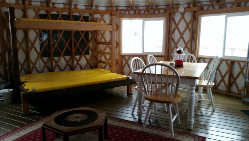 Interior of a yurt featuring a yellow mattress, dining table with chairs, and large windows.
