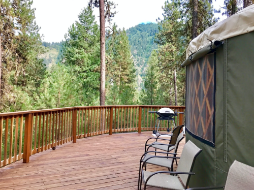 A wooden deck surrounded by tall trees, featuring a grill and chairs, with mountains in the background.
