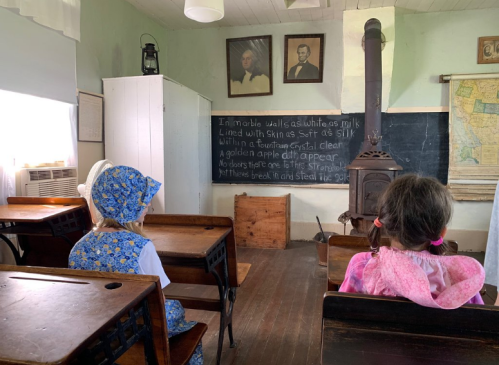 Two children in vintage clothing sit in a historic classroom, facing a chalkboard with handwritten text and old portraits on the wall.