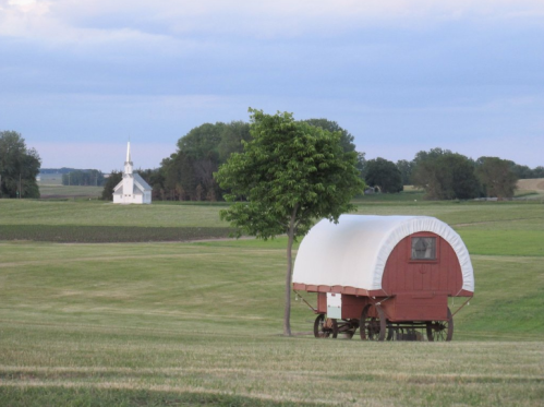 A red covered wagon sits in a grassy field with a white church and trees in the background under a cloudy sky.