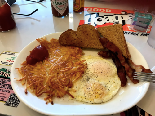 A plate of breakfast featuring two fried eggs, crispy bacon, hash browns, toast, and ketchup on the side.