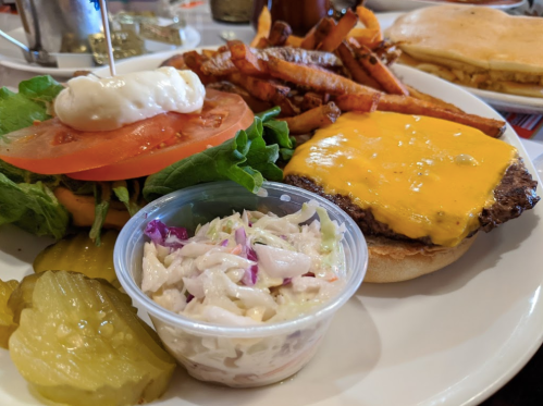 A plate featuring a cheeseburger with lettuce, tomato, and pickles, alongside sweet potato fries and coleslaw.