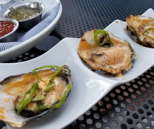 Grilled oysters topped with green onions, served on a white plate with dipping sauces in the background.