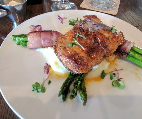 A plated dish featuring seasoned fish, asparagus wrapped in bacon, and creamy mashed potatoes, garnished with microgreens.