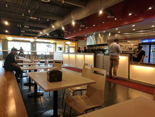 A modern restaurant interior with tables, a counter, and customers ordering at the front. Bright lighting and a casual atmosphere.
