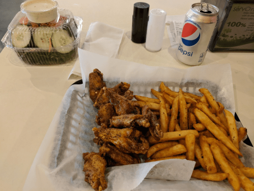 A tray with seasoned chicken wings and fries, alongside a salad and a can of Pepsi on a table.