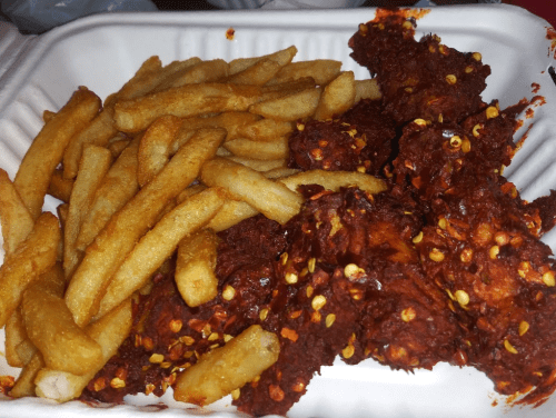 A takeout container with crispy fries and spicy, red-coated chicken pieces sprinkled with chili flakes.