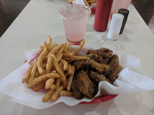 A red basket with crispy fried chicken wings and golden French fries, accompanied by a pink drink and condiments.