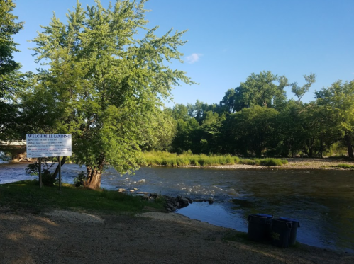 A serene river scene with trees, a sign for Welch Mill Landing, and two bins near the water's edge.