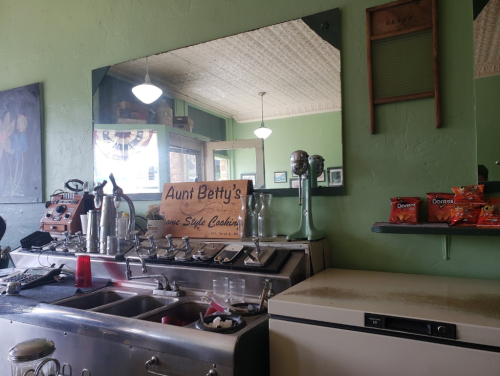 A retro diner interior featuring a mirrored wall, vintage appliances, and a sign for "Aunt Betty's Home Style Cookin'."