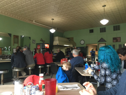 A cozy diner scene with patrons at tables and the counter, featuring green walls and vintage decor.
