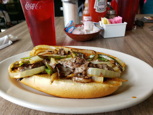 A loaded sandwich with beef, grilled onions, and peppers on a toasted bun, served with drinks in the background.