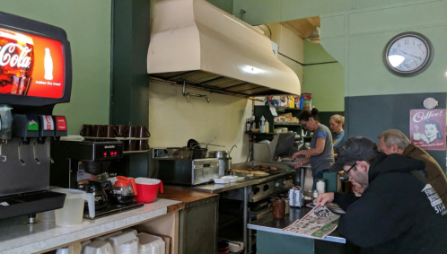 A cozy diner kitchen with a cook at the grill, customers seated, and a soda machine in the foreground.