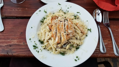A plate of pasta topped with grilled chicken, garnished with parsley and grated cheese, on a wooden table.