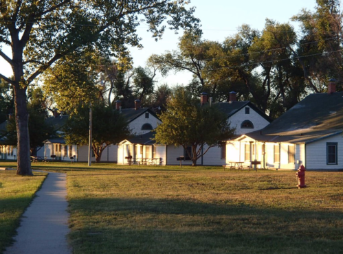 Sunlit row of white houses with porches, surrounded by trees and a grassy area, in a peaceful neighborhood setting.