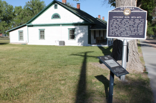 Historic building with a green roof and white siding, featuring informational signs in front on a sunny day.