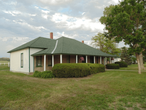 A green-roofed house with a porch, surrounded by manicured lawns and trees under a cloudy sky.