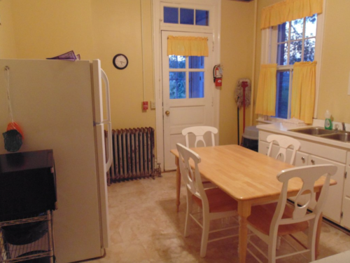 A bright kitchen with yellow curtains, a wooden table, white chairs, a refrigerator, and a door leading outside.