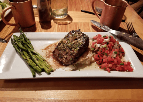 A plate featuring a grilled steak on mashed potatoes, asparagus, and fresh tomato salsa, with copper mugs in the background.