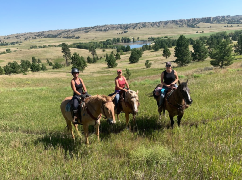 Three women on horseback in a grassy field with rolling hills and a lake in the background on a sunny day.