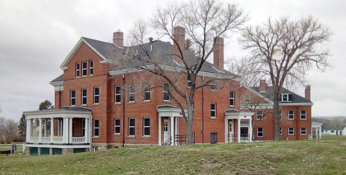 Historic red brick building with multiple chimneys, large windows, and a porch, surrounded by bare trees and grass.