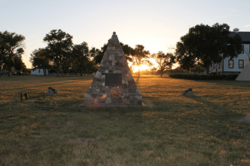 A stone monument stands in a grassy field at sunset, with trees and a building in the background.