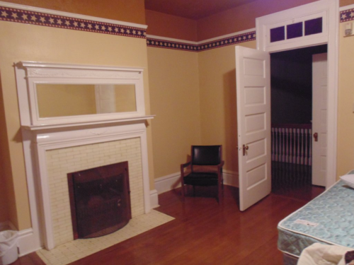 A cozy room with a fireplace, a chair, and an open door leading to a dark hallway. Walls are painted yellow with a decorative border.