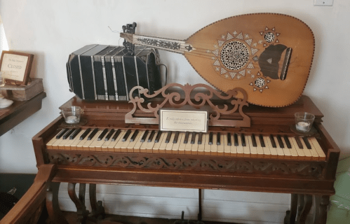 A wooden piano with decorative carvings, accompanied by an accordion and a stringed instrument, displayed in a cozy setting.