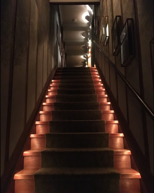 A dimly lit staircase with illuminated steps leading up to a hallway adorned with framed pictures.