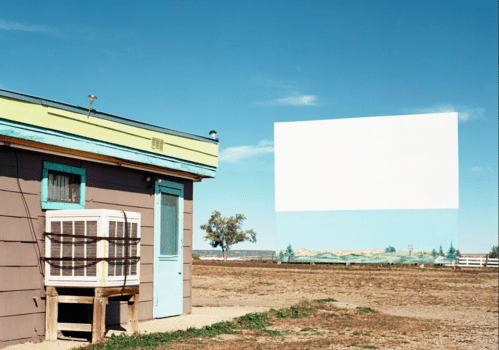 A small, rustic building with an air conditioner beside a large, blank outdoor screen against a clear blue sky.
