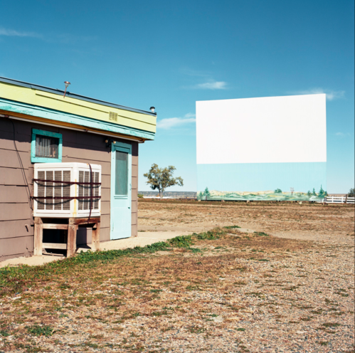 A rustic building with a turquoise door beside a large, blank movie screen in an open, barren landscape.