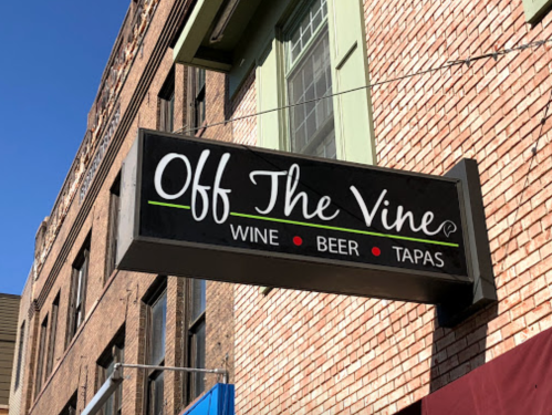 Sign for "Off The Vine" featuring wine, beer, and tapas, mounted on a brick building under a clear blue sky.