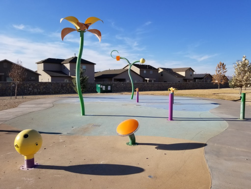 Colorful water play area with flower-shaped features and spray nozzles, set against a clear blue sky and houses in the background.
