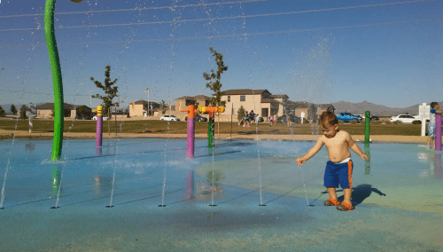 A young child plays in a splash pad, surrounded by colorful water features and houses in the background.