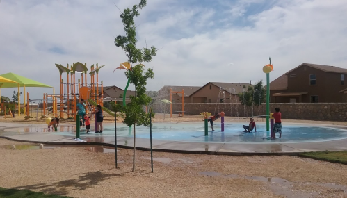 Children playing in a splash pad area with colorful play structures and a few trees under a partly cloudy sky.
