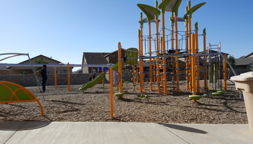 A colorful playground with climbing structures and swings, set against a clear blue sky and residential buildings.