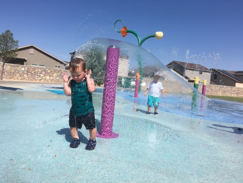 A toddler plays in a splash pad, water spraying around him, while another child stands in the background.
