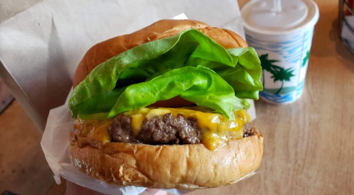 A close-up of a juicy cheeseburger with lettuce and a slice of cheese, held in a hand, with a drink in the background.