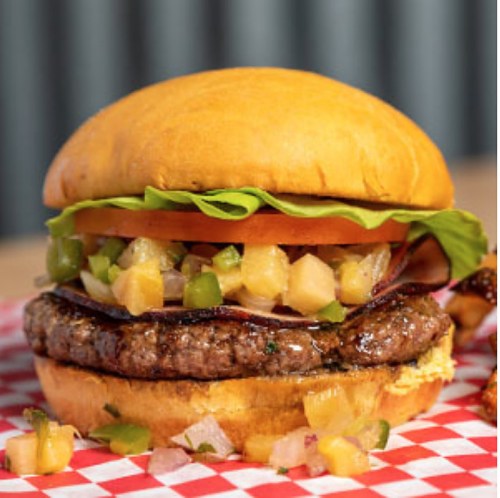 A juicy burger with lettuce, tomato, onions, green peppers, and pineapple salsa on a toasted bun, served on a checkered paper.