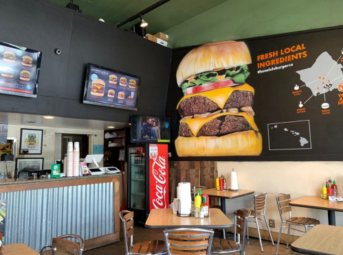 Interior of a burger restaurant featuring a large burger mural, menu screens, and tables with condiments.