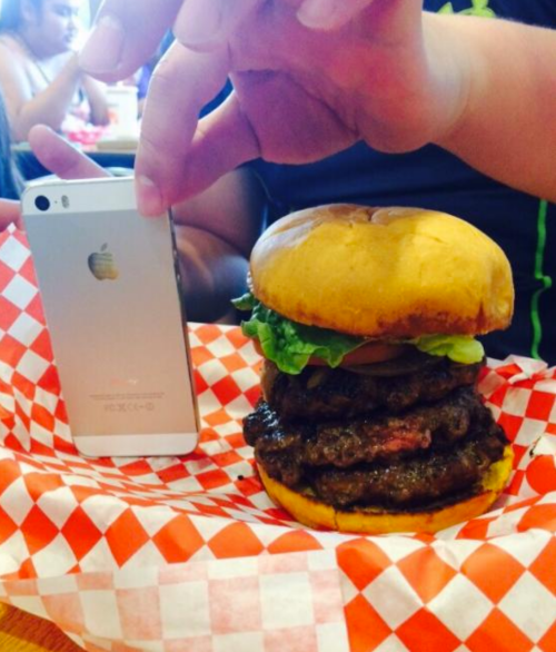 A hand holds a smartphone next to a large burger with three patties, lettuce, and tomato on a checkered paper.