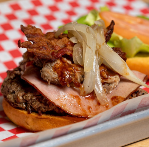 A hearty burger topped with bacon, grilled onions, and ham, served on a bun with lettuce and tomato on the side.