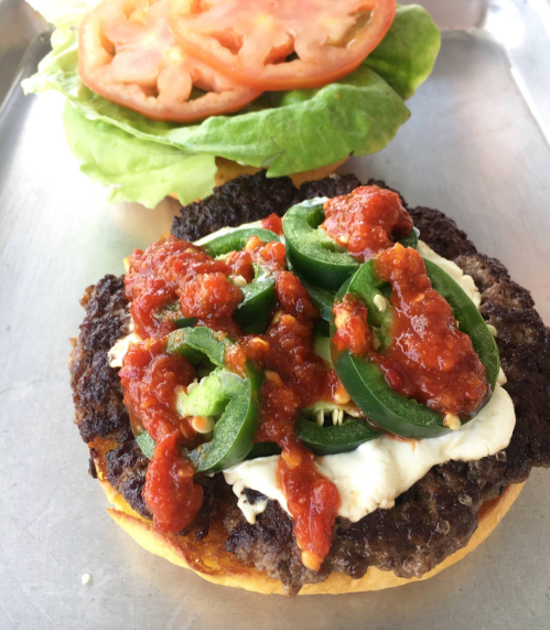 A close-up of a burger patty topped with jalapeños, salsa, and creamy sauce, with lettuce and tomato on the side.