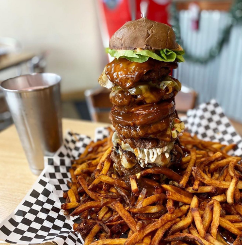 A towering burger with multiple patties and toppings sits atop a large serving of crispy fries on a checkered plate.