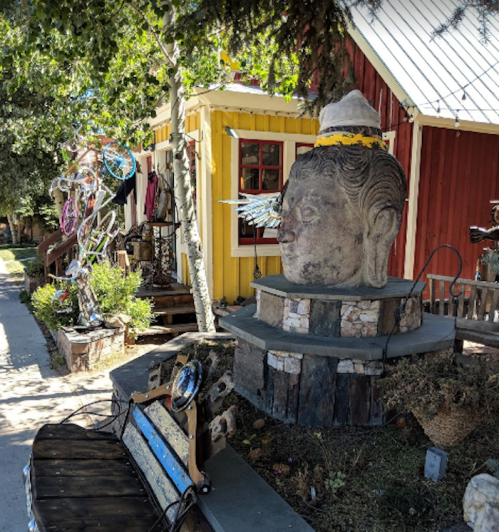A large stone Buddha head sculpture sits on a stone base in front of a colorful house with artistic decorations.