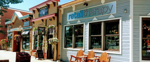 A charming storefront with the sign "retail therapy," featuring outdoor seating and decorative flowers.