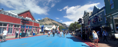 A vibrant street scene with a blue road, lined by colorful buildings and people, set against a mountain backdrop.
