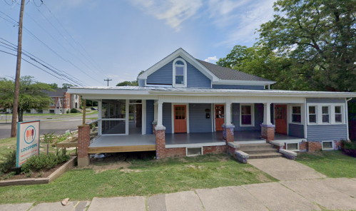 A blue and gray house with a porch, featuring orange doors and a sign, surrounded by greenery and a sidewalk.