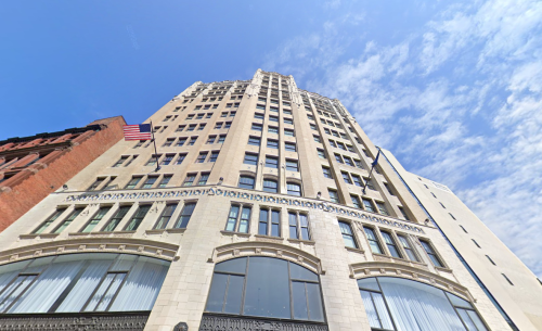 A tall historic building with a flag, viewed from below against a blue sky with clouds.