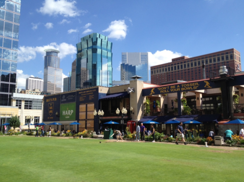 A vibrant urban scene with green grass, buildings, and people enjoying a sunny day at a bowling venue.